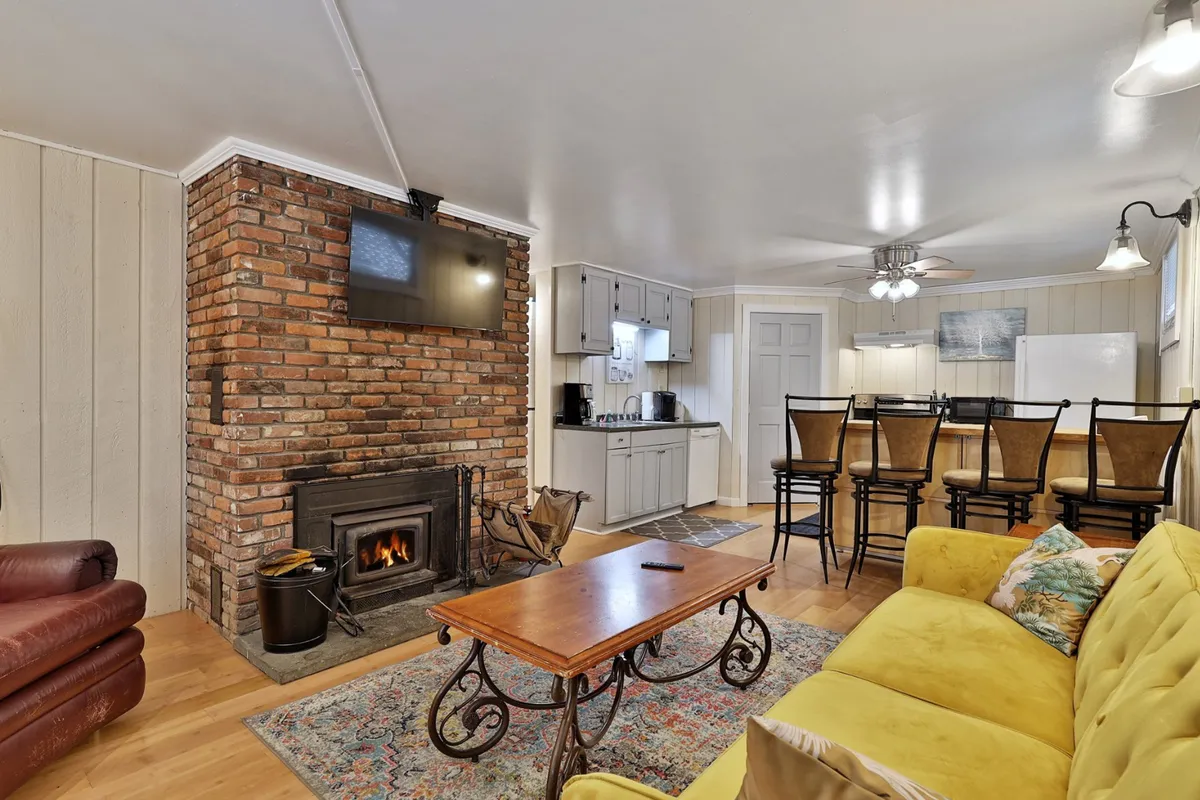 The image showcases a warm and inviting living space centered around a rustic brick fireplace, with a cheerful yellow sofa and a wooden coffee table inviting relaxation. Bar stools line a nearby counter, hinting at a space that's perfect for gathering, enjoying hot cocoa after a day on the slopes, or sharing stories by the fire. The overall mood is cozy and welcoming, ideal for unwinding with friends or family after a day in the Vermont snow.