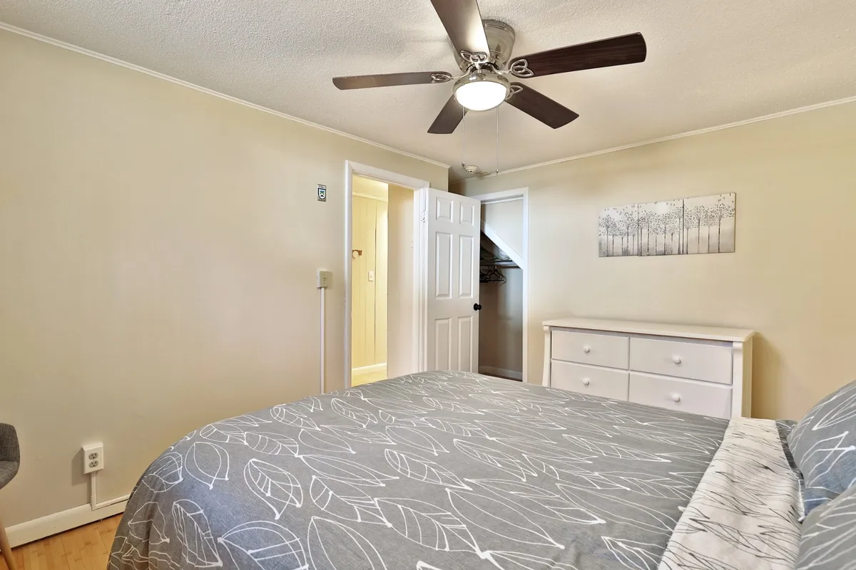 A cozy bedroom scene showing a queen-sized bed with patterned bedding, a white dresser for storage, and a ceiling fan. The space feels both inviting and functional, highlighting comfort and practicality.