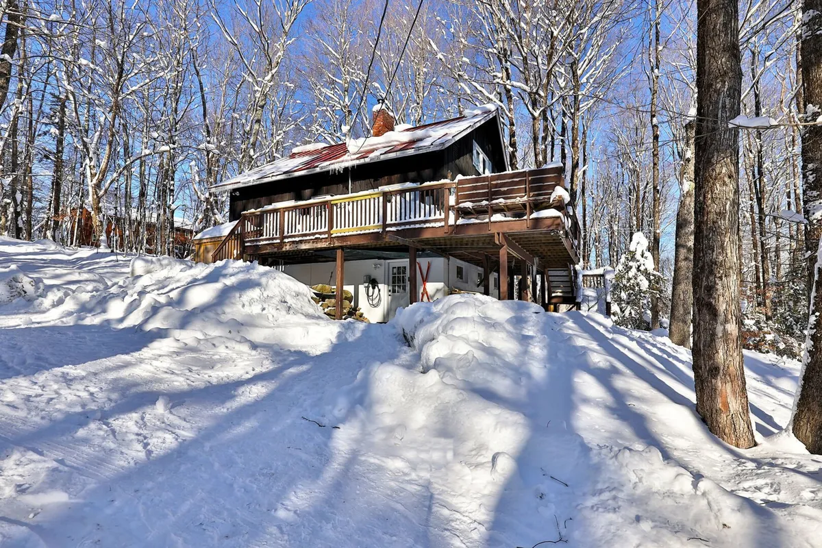 A snowy landscape with a wooden deck in the foreground, surrounded by a serene tree line. The entire scene is blanketed in fresh snow, evoking the quintessential winter getaway atmosphere. The setting suggests easy access to winter sports and cozy gatherings after a day outside.