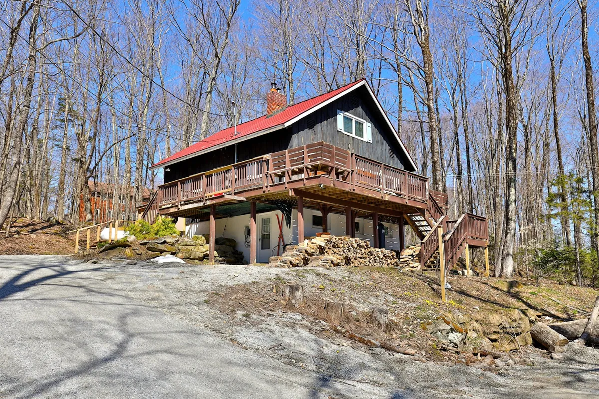 A cozy wooden deck with a red roof, surrounded by trees and a stone pathway, featuring a firewood stack that hints at warmth and comfort. The setting suggests both adventure in the surrounding nature and a welcoming return to the property.