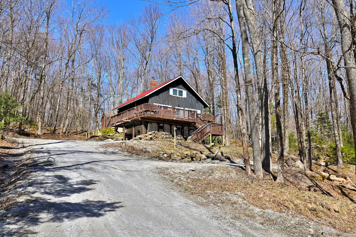 A welcoming view of the house with its distinctive red roof, expansive driveway for easy parking, and lush trees providing a natural, secure setting. The clear view of the driveway highlights the accessible entryway and space for guest vehicles.