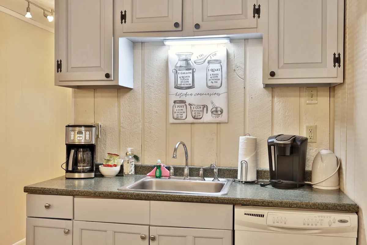 The image emphasizes the kitchen sink area, featuring a coffee maker and decorative wall art. There's a practical yet cozy vibe, with hints of daily rituals like brewing coffee, cleaning up, and sharing conversations over a warm drink.