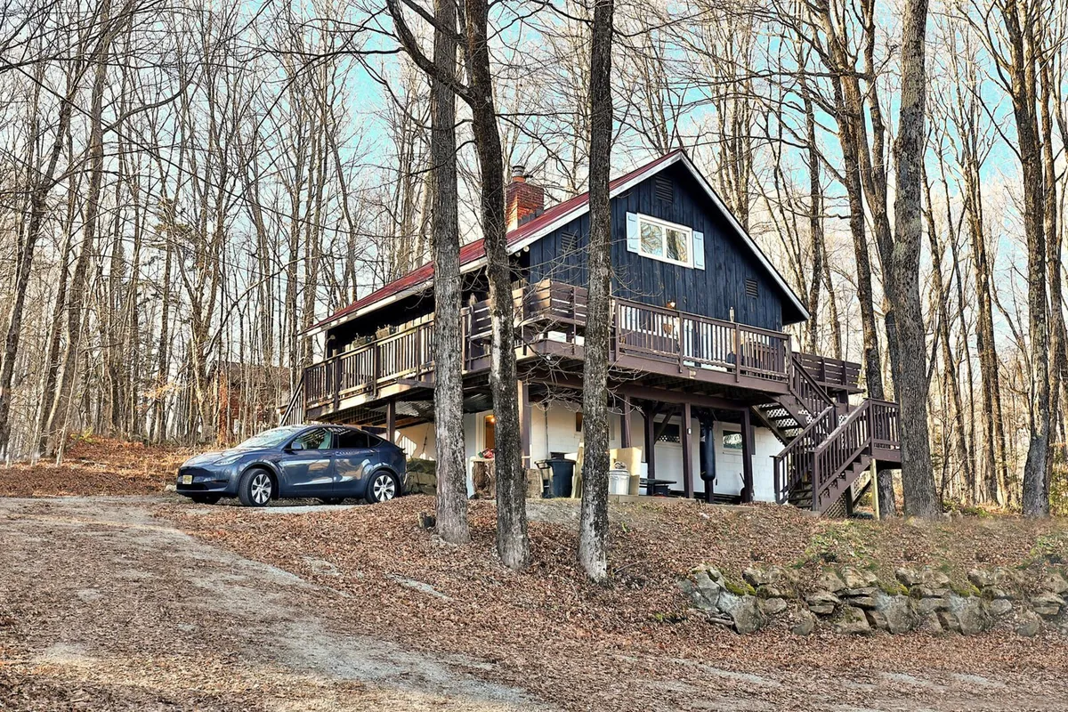 This image displays the exterior of the house, showcasing the deck, surrounding trees, and driveway. The scene suggests accessibility to nature and outdoor relaxation spaces, with ample room for exploring or spending time with pets. The peaceful setting hints at direct proximity to trails and the Vermont outdoors.