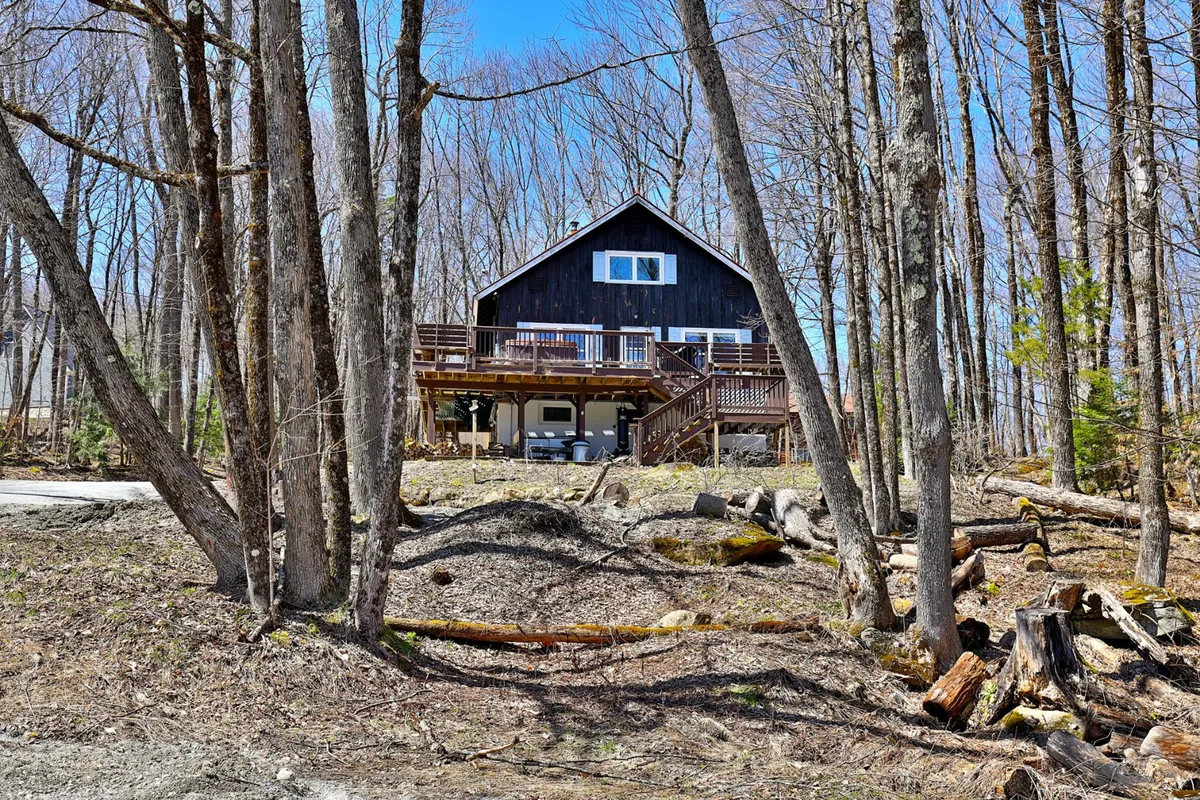 The exterior of the home is framed by tall trees, highlighting the house, deck, and natural surroundings. The atmosphere is tranquil, emphasizing relaxation, outdoor gatherings, and a close relationship with the wooded landscape.
