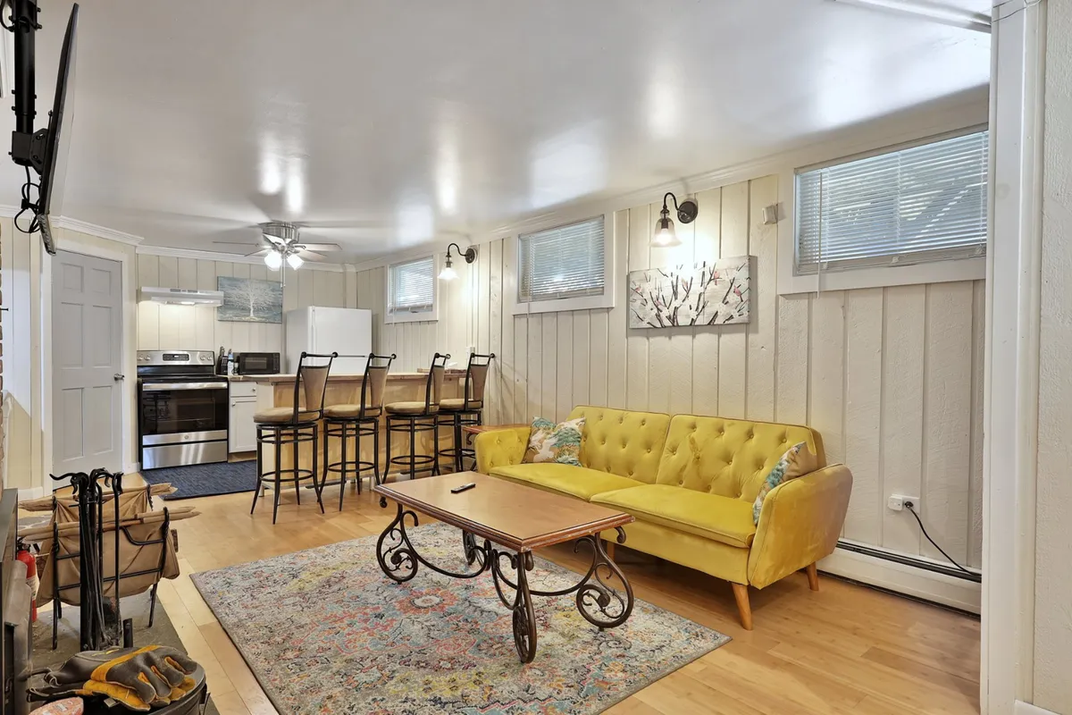 A warm, welcoming living area featuring a bright yellow sofa, wooden coffee table, and a modern kitchen in the background. Bar stools line the kitchen counter, and stainless appliances are visible, suggesting a comfortable space for gathering after outdoor adventures.